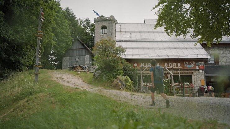 Ein junger Wanderer geht auf einer Schotterstraße an einer Berghütte vorbei in Richtung Turm