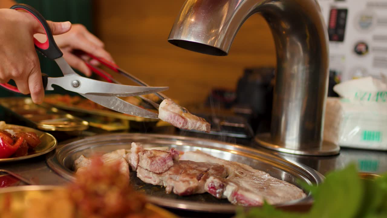 Person uses scissors to cut grilled meat on tabletop grill in brightly lit restaurant setting