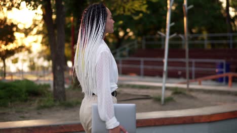 Confident-walk-of-and-elegant-girl-in-white-blouse-and-pants---young-girl-walks-through-the-park-with-a-laptop-in-her-hand.-Side