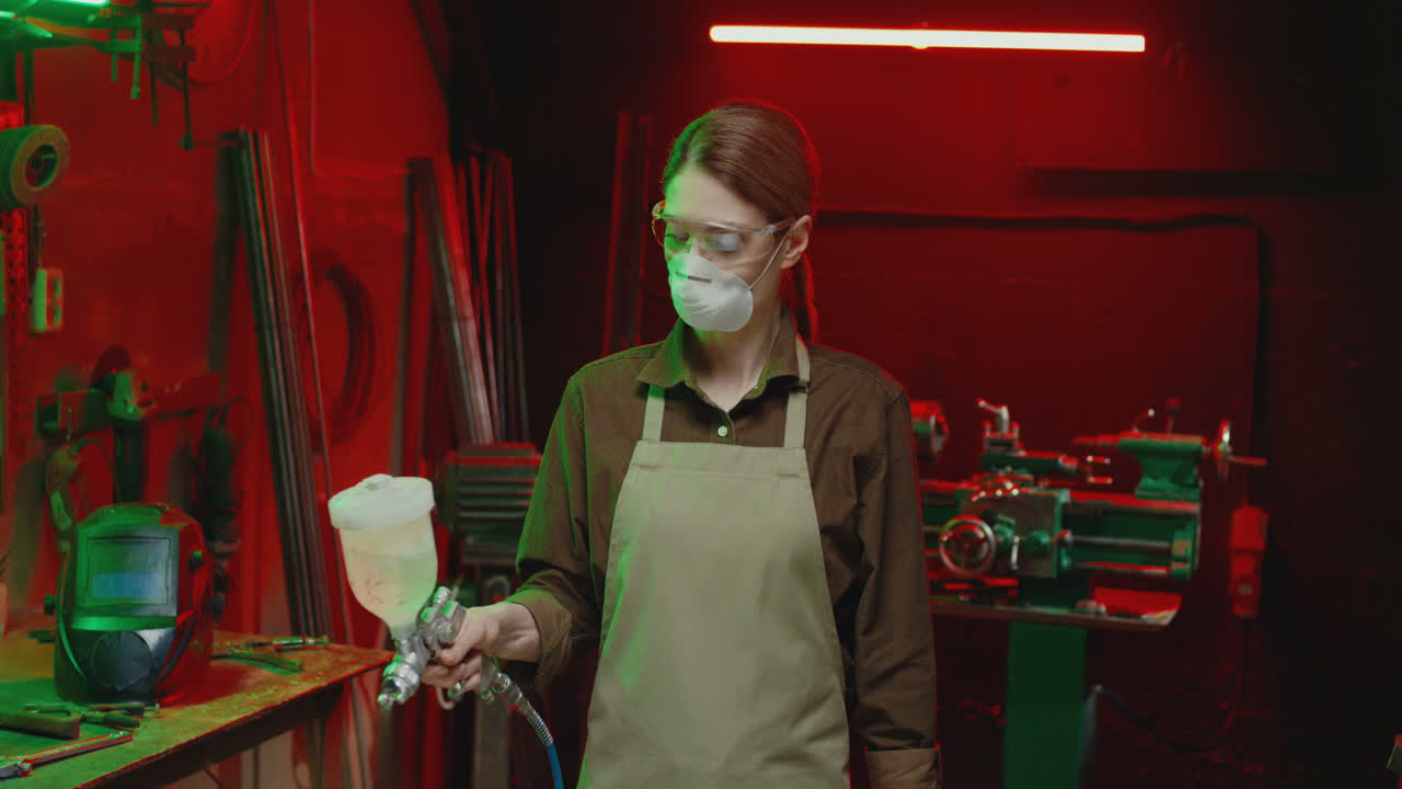 кавказская женщина-сварщик в защитных очках, респираторной маске и фартуке работает с пульверизатором в мастерской