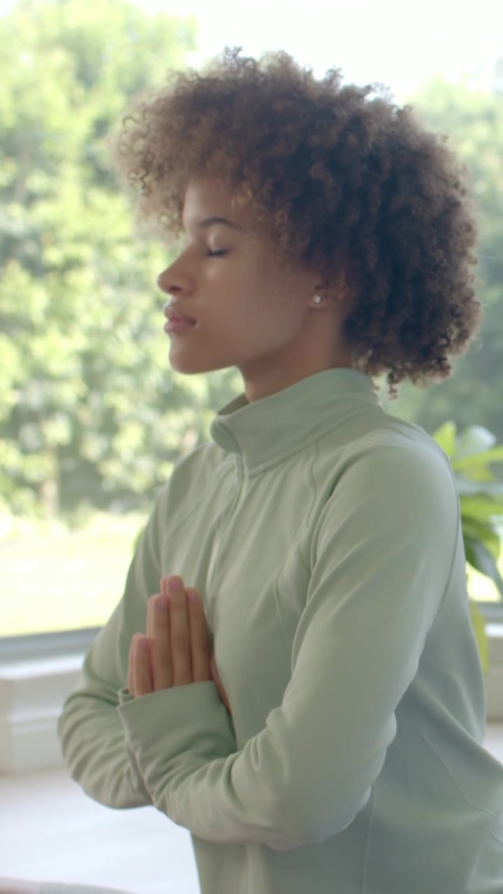 Vertical video: Settling woman in light green top pressing palms, meditating by window with plant