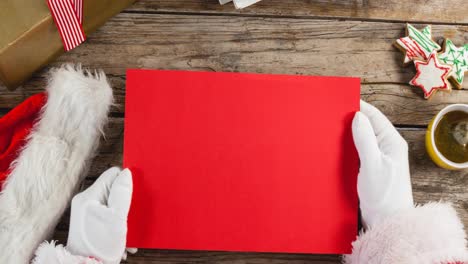 animation of hands of santa claus holding red christmas card with copy space over wooden background