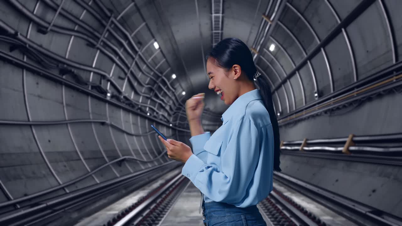Side View Of Asian Female Professional Worker With Her Smartphone In Underground Subway Tunnel, She Raises Her Fist Up With Screaming Goal After Check On The Smartphone
