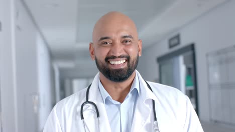 portrait of happy biracial male doctor wearing lab coat, smiling in corridor, slow motion