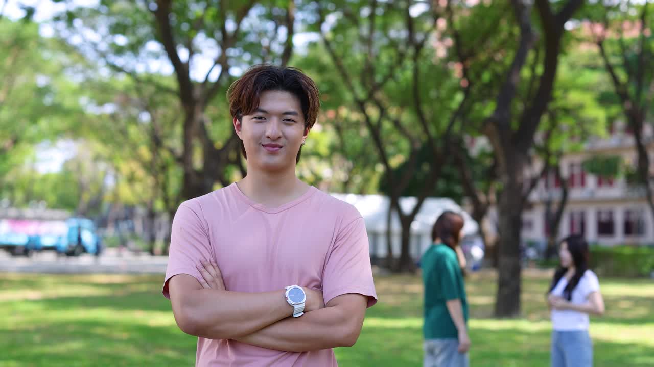 ピンクのシャツを着た若いタイ人男性が、大学の敷地内で自信に満ちた笑顔を見せている。