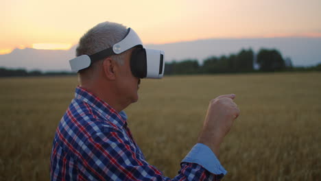 an elderly male farmer uses gestures with his hands standing in a field wearing virtual reality glasses. use vr glasses in agriculture on a field with wheat.