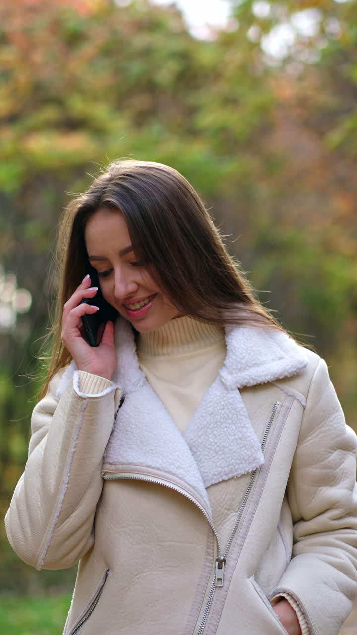 Beautiful dark-haired lady in white jacket walking slowly by the autumn wood. Attractive woman speaks on the phone continuing her walk. Vertical video