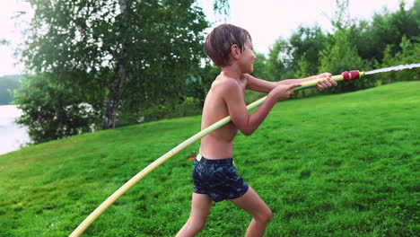 Junge-In-Sommerbadehose-Gießt-Wasser-Auf-Seinen-Jüngeren-Bruder,-Der-Sich-Im-Park-Auf-Dem-Gras-In-Der-Nähe-Des-Sees-Vergnügt