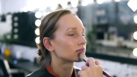 Hand-of-make-up-artist-applying-tone-on-a-model's-face-using-a-big-brush.-Beautiful-caucasian-blonde-woman.-Blurred-make-up