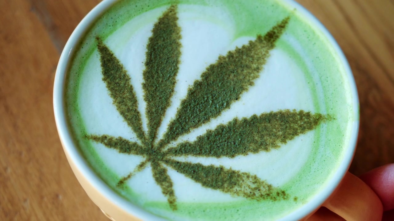 Vista dall'alto di un matcha latte in una tazza di ceramica, decorato con tè verde in polvere che forma un motivo a foglia che ricorda una foglia di cannabis.