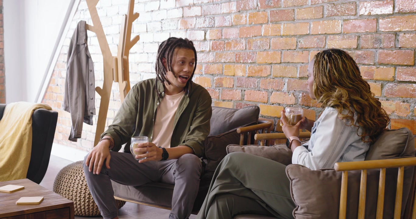 African American couple sitting on couch, man smiling, starting chat and raising clear glass mugs