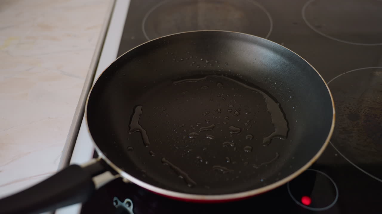 Close up of frying pan with oil heating on electric stove in bright kitchen, showing drops of oil spread inside pan with focus on cooking surface and kitchen counter in background