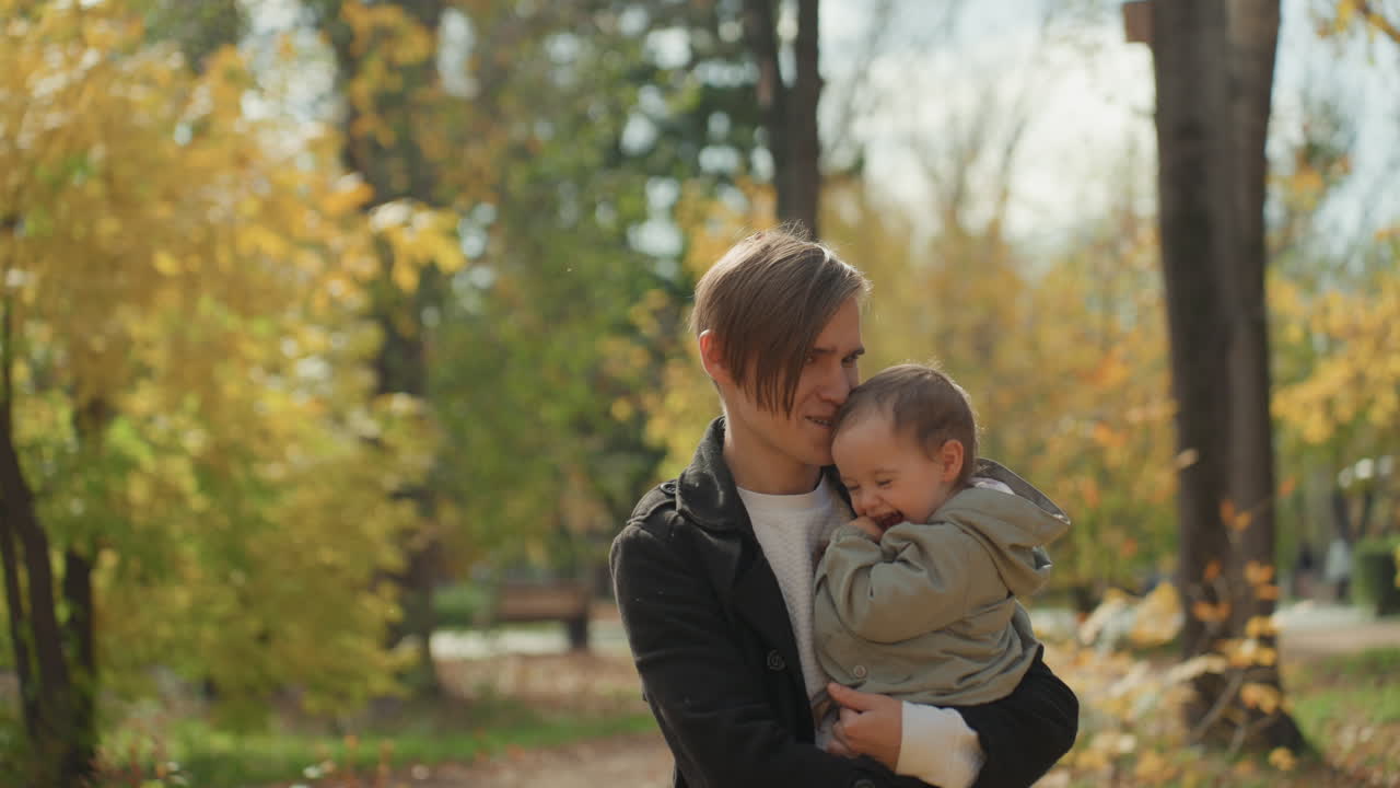 Padre caucasico tiene in braccio il bambino mentre il partner lancia foglie nel parco, una cattura spontanea di un giovane papà caucasico che sorride con il figlio mentre la mano del partner scaglia il fogliame dorato, una calda scena di una gita in famiglia.