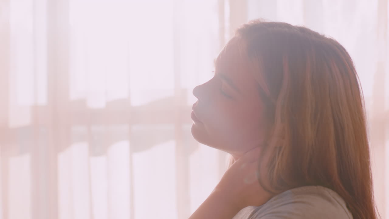 Close up side view of young woman holding back of neck while gently tilting head under soft sunlight through curtains, conveying physical discomfort from tension or fatigue