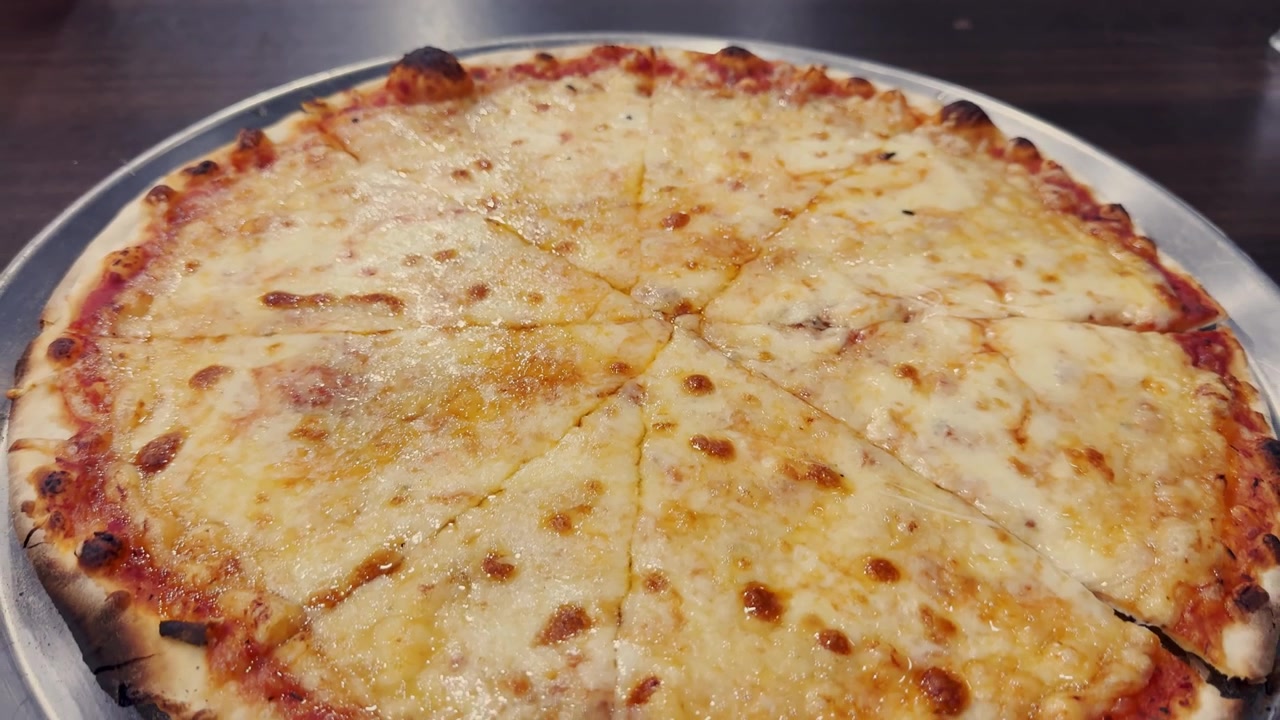 Primo piano di una pizza al formaggio a fette su un vassoio di metallo, con mozzarella fusa, salsa di pomodoro, crosta dorata e un'apparecchiatura informale da ristorante.