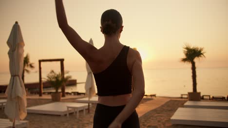 a brunette girl with tied hair in a black sports summer uniform holds her hands behind her back and does stretching on a sunny beach in the morning