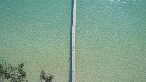 aéreo, de arriba hacia abajo, ojo de pájaro, un dron disparó sobre una mujer caminando y un hombre corriendo en un muelle de madera, mientras la gente nadaba en el mar turquesa, en una playa paradisíaca, en un día soleado, en koh kood, tailandia, asia