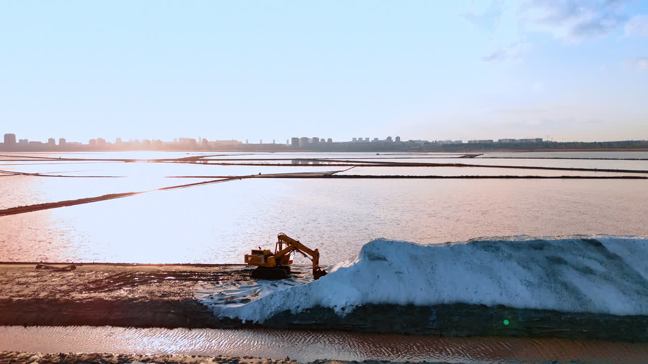 Escavatore industriale al lavoro in una salina al tramonto. Macchinari pesanti che spostano un cumulo di sale bianco in una salina costiera con il riflesso del sole sull'acqua.