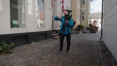 wide shot of camera man in blue jacket setting up cinema camera on a tripod on a small town main street