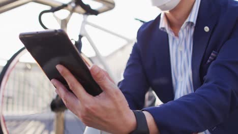 Asiatischer-Mann-Mit-Gesichtsmaske-Und-Digitalem-Tablet,-Während-Er-Auf-Der-Treppe-Im-Firmenpark-Sitzt