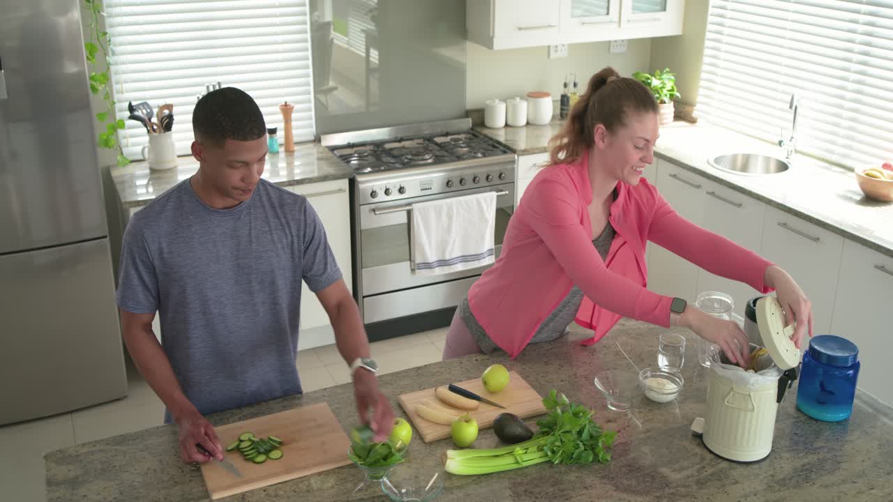 Casal com homem cortando pepino e mulher enchendo liquidificador enquanto preparam suco na ilha da cozinha