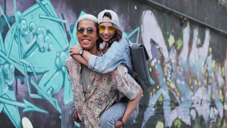 young boy giving her girlfriend a piggyback and looking at the camera near a graffity wall in the street