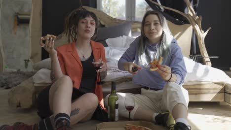 front view of two happy girls drinking wine and eating pizza while sitting on the floor and watching tv