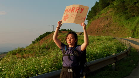 young solo female asiatic traveller hold a sing on cardboard while hitching on road side during a backpacking adventures trekking trip