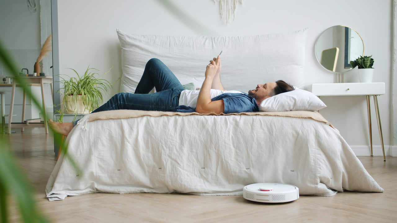 Man relaxing on bed with smartphone, robot vacuum cleaner