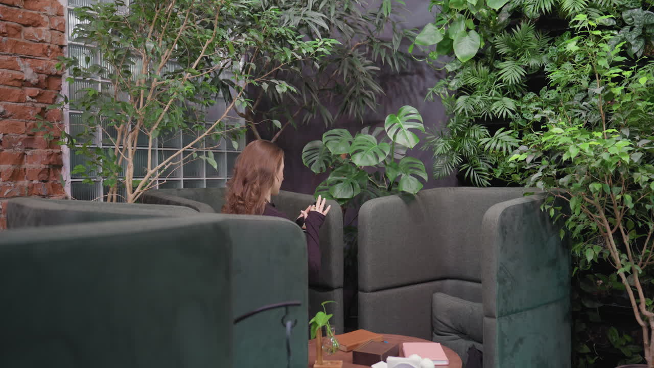 Pretty woman holding phone while standing between cushioned chairs in lush indoor setting filled with various green plants, leafy decoration, brick wall, and glass panels in background