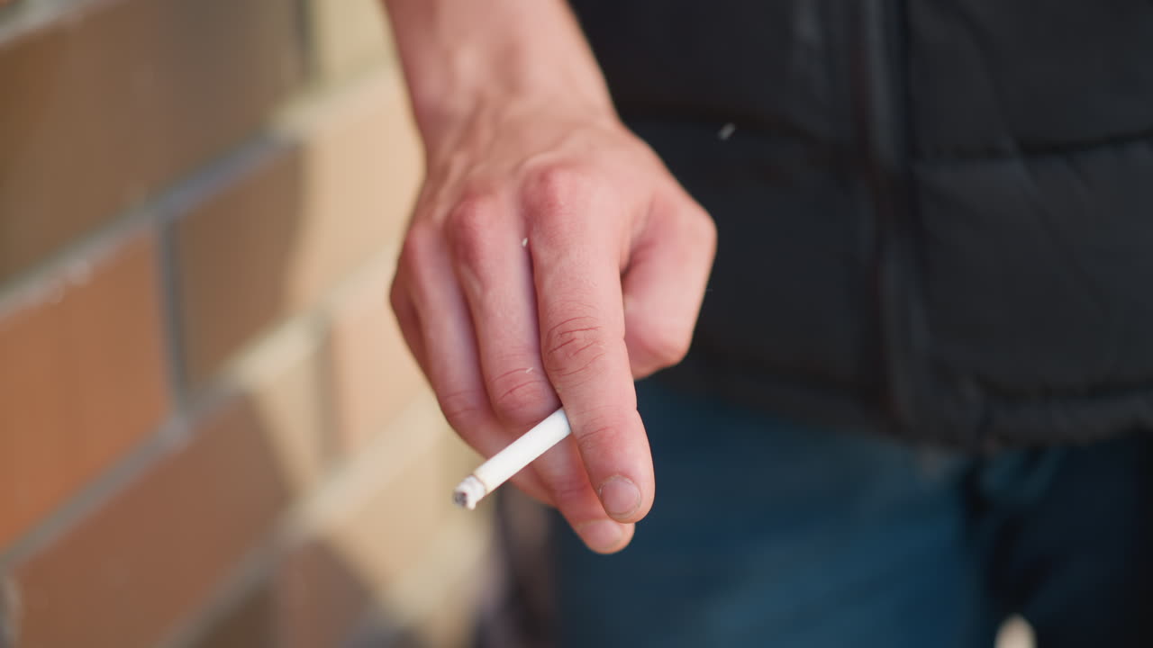 partial view of smoker flicking cigarette as ash drops while sunlight casts animated shadow across nearby brick wall, capturing subtle movement and casual gesture