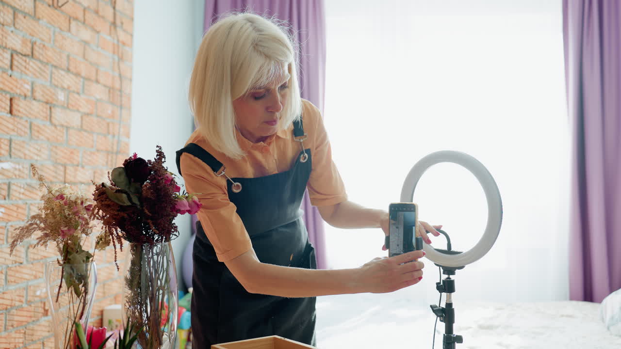 Woman florist in apron adjusting smartphone on tripod with ring light preparing to record floral tutorial indoors, surrounded by vases with flowers and creative workspace background with toys and curtains
