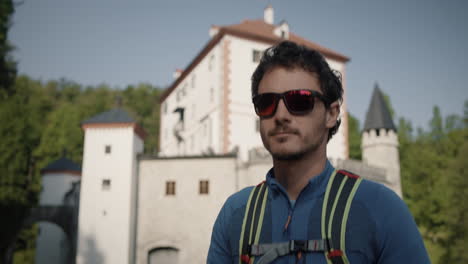 Camera-tracking-hiker-walking-infront-the-castle-Snežnik-in-a-sunny-day,-wearing-sunglasses-and-light-green-backpack
