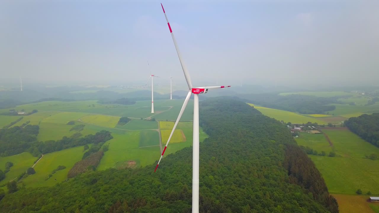 A breathtaking view of a wind turbine in the vast rural landscape of Mörsdorf, Germany. The turbine stands tall over fields and forests, harnessing the wind to generate sustainable electricity.