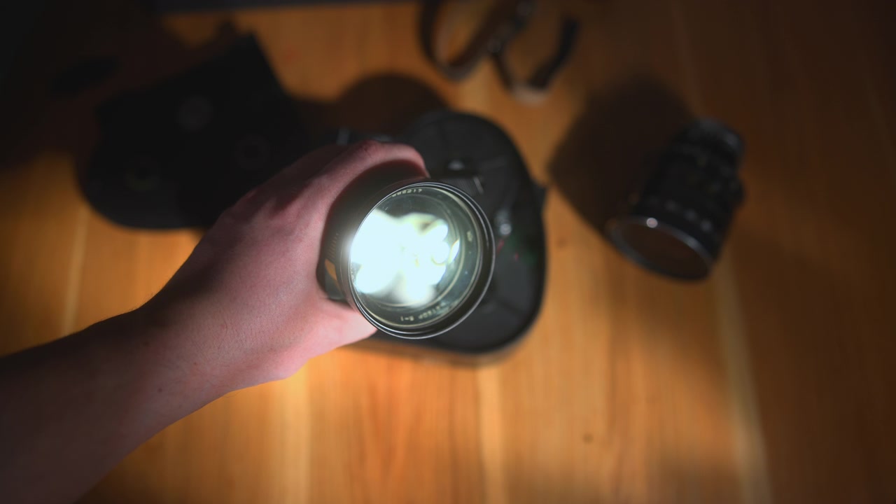 Uma mão segura uma lanterna brilhante apontada para o observador acima de uma mesa de madeira, enquanto lentes de câmera desfocadas e equipamentos escuros estão no fundo quente e com pouca luz.