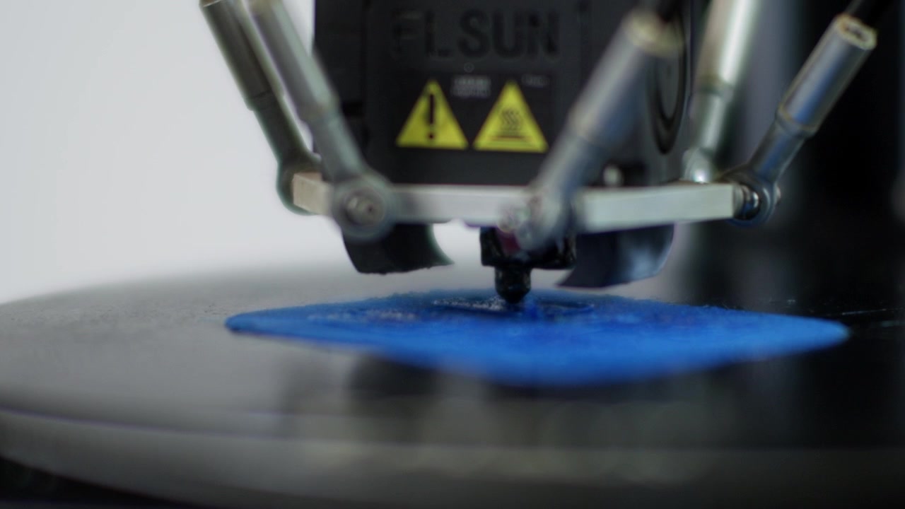 Extreme macro shot focusing on the nozzle of a delta 3D printer as it extrudes a layer of blue plastic filament onto the build plate.