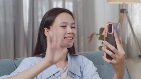 close up of asian teen girl having video call on smartphone while sitting on sofa in the living room at home. waving hands, smiling, and speaking