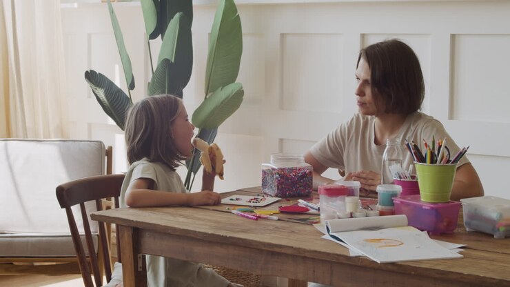 A Happy Mother Offers A Banana To Her Pretty Blonde Daughter As They Play Together With Colored Beads