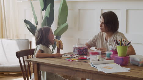 una madre feliz le ofrece un plátano a su linda hija rubia mientras juegan juntas con cuentas de colores