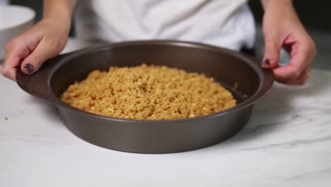 Vista-De-Cerca-De-Las-Manos-Femeninas-Agitando-Trozos-De-Masa-En-Una-Fuente-Redonda-Para-Hornear.-Preparación-De-Tarta-De-Queso.-Repostería-Casera.-Tiro-En-Cámara-Lenta