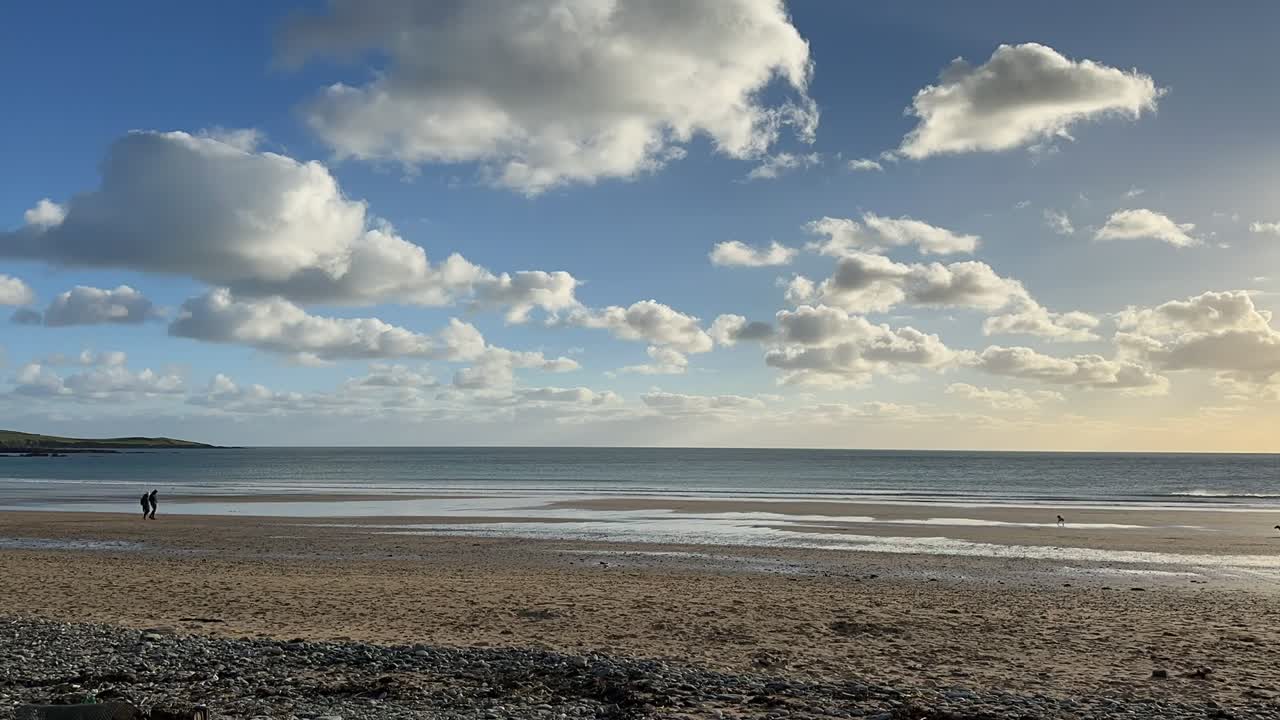 Nuvole bianche e soffici che passano in un cielo invernale azzurro e soleggiato, mentre persone camminano su una spiaggia sabbiosa in un video in time-lapse.