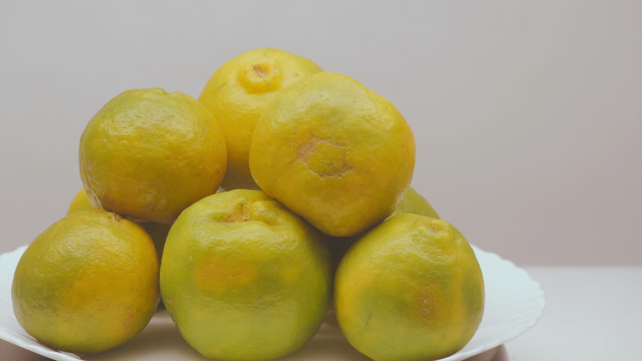 A neat stack of ripe oranges slowly rotates on a white display plate, revealing their mixed yellow–green tones and natural textures under soft studio lighting