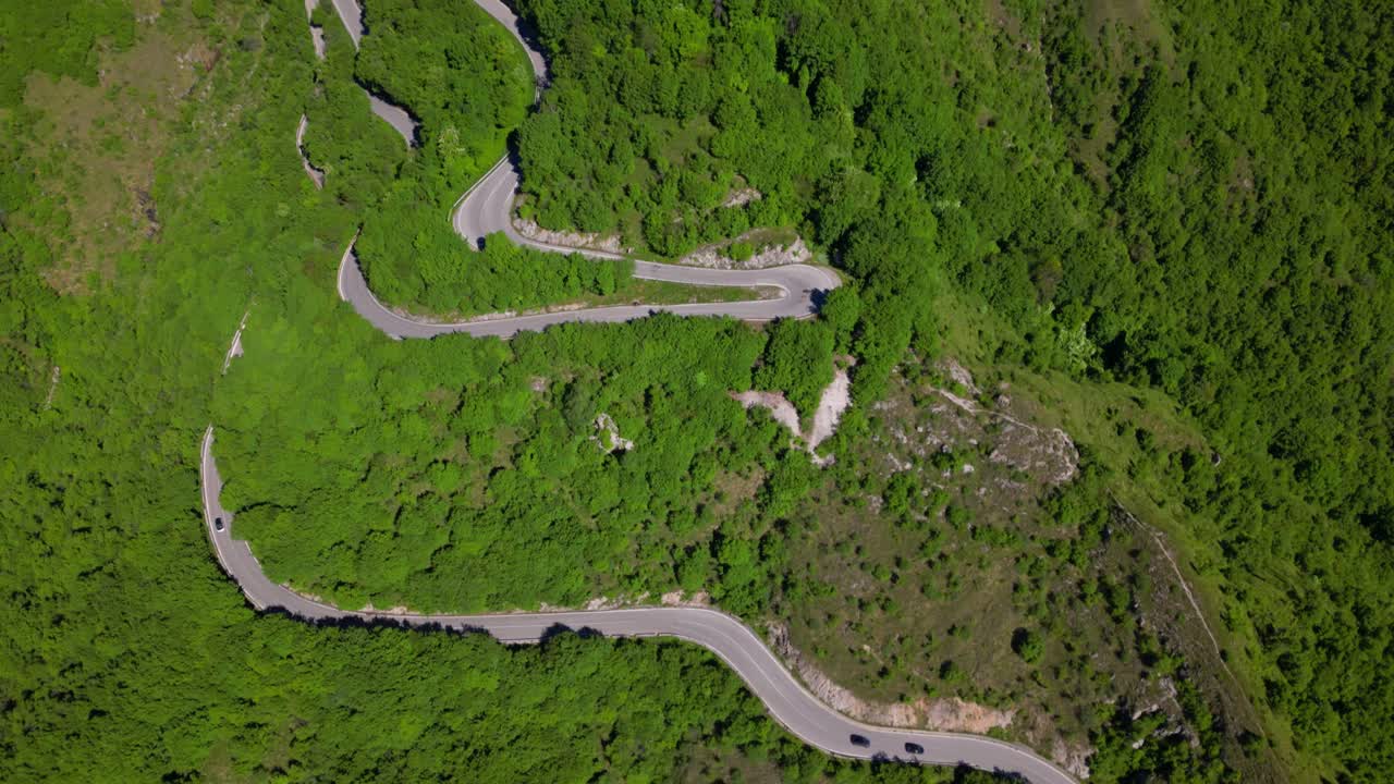 Wide top-down aerial shot of multiple switchbacks cutting through the forested mountain slope with sparse traffic. Shot at Selvino, Italy (Selvino, Italia)