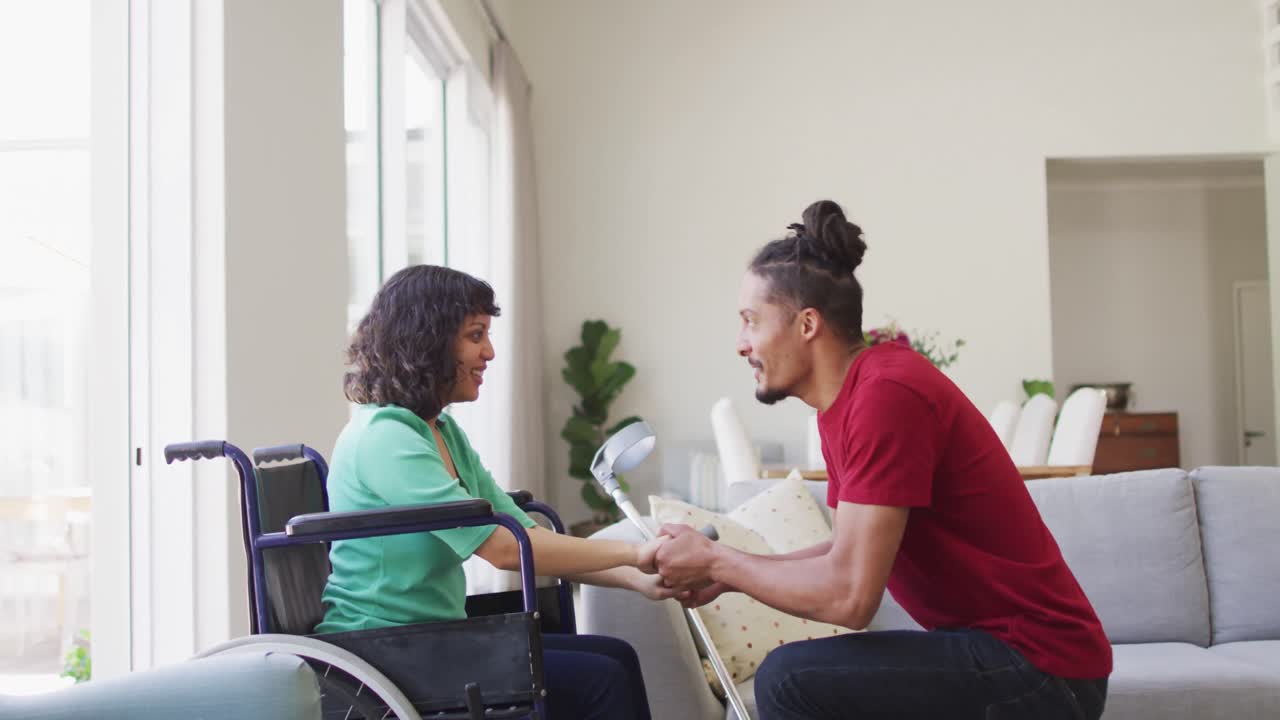 mujer biracial feliz en silla de ruedas y compañero masculino sonriente sentado de la mano en la sala de estar