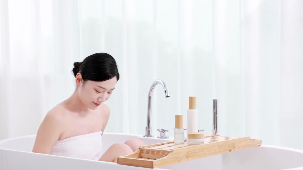 シャワー後、バスタブに座ってリラックスしながら、タオルに包まれた若い女性が手に保湿クリームやボディローションを塗っている。スキンケア、セルフラブ、ボディケア、自宅スパトリートメント。