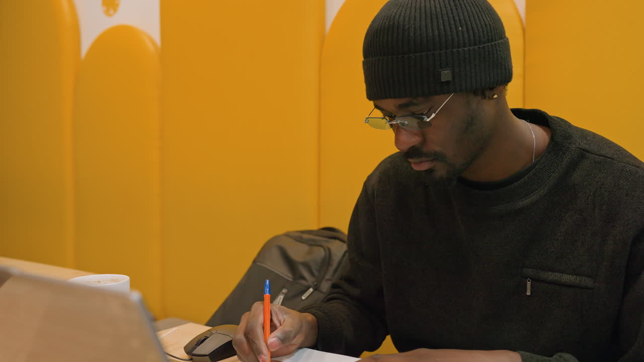 Designer negro esboçando na mesa do laptop, criando desenhos conceituais no papel, canetas coloridas e óculos na mesa, fundo de divisória amarela, ambiente de estúdio colaborativo, brainstorming freelancer.