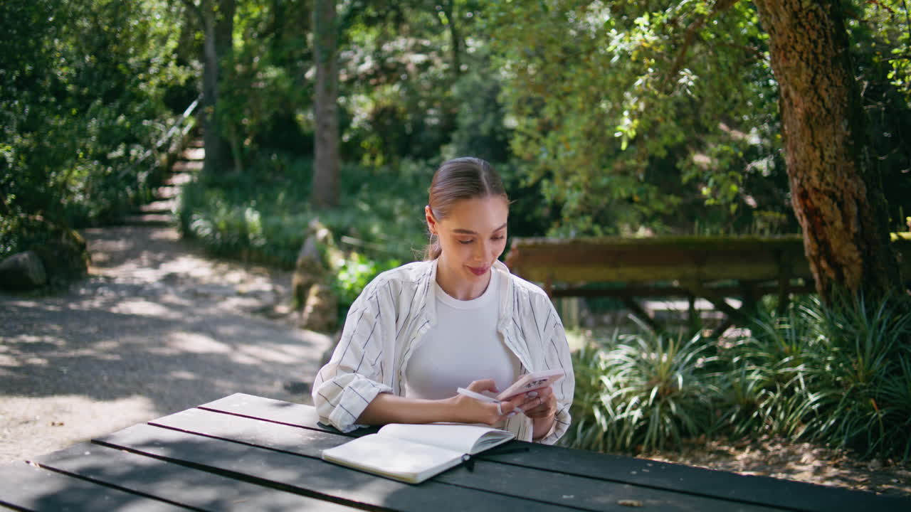 Donna felice che legge un messaggio sul cellulare, seduta al tavolo del parco con un libro, in primo piano.
