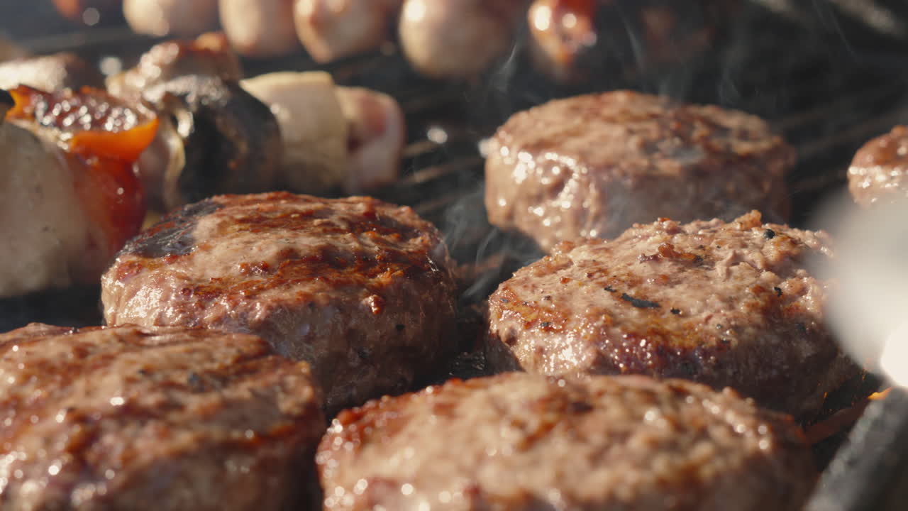 Hamburger di manzo affumicati e cotti alla fiamma su griglia barbecue sfrigolante con spiedini e salsicce sullo sfondo