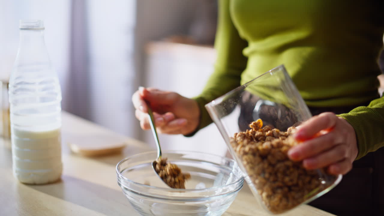 Primo piano delle mani di una casalinga che mettono la granola in una ciotola di vetro. Donna sconosciuta che cucina.