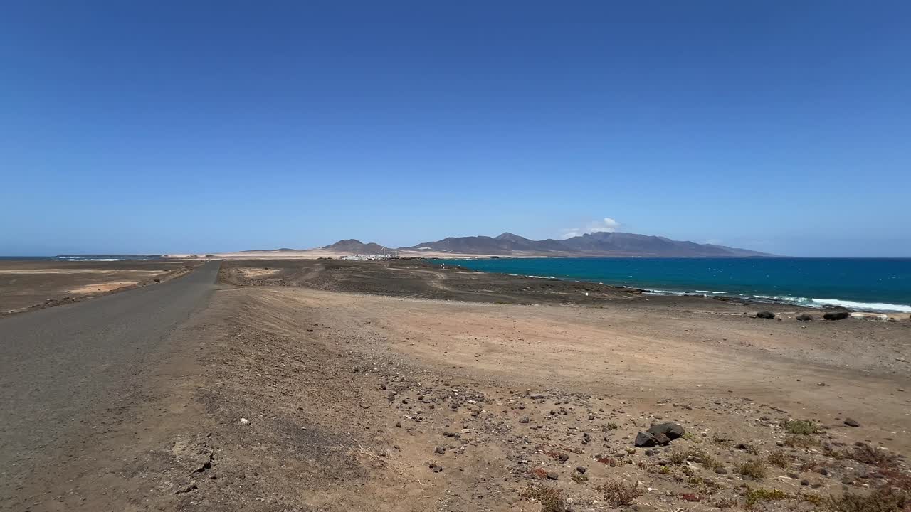 POV driving along a remote coastal dirt road in the Jandia peninsula, with the arid landscape meeting the blue Atlantic Ocean. Fuerteventura, Spain